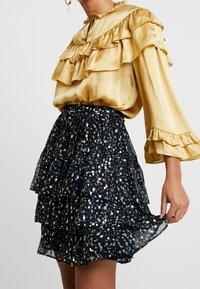 Blusa dorata con ruches e maniche lunghe abbinata a una gonna nera con tessuto a strati e un motivo astratto colorato.