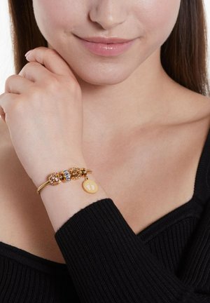 Gouden armband met kleurrijke kralen, waaronder roze en blauwe accenten, met een ronde charme waarop voetafdrukken zijn afgebeeld. Draagt op een pols, met een gestructureerde zwarte top.