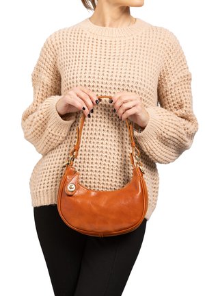 Braune Ledertasche mit einer geschwungenen Form, kurzen Griff und goldenen Hardware-Details, die von einer Person getragen wird, die einen beigen Strickpullover trägt.