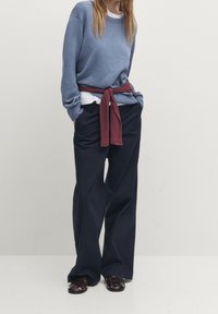 Blue knitted sweater layered over a white shirt, with a red tie belt around the waist, paired with loose navy trousers and brown sandals.