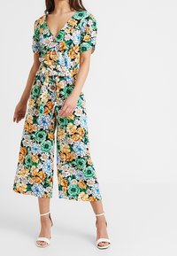 Kwiatowy kombinezon z dekoltem w kształcie litery V, krótkimi rękawami i paskiem w talii. Charakteryzuje się odważnym połączeniem pomarańczowych, zielonych i niebieskich kwiatów na czarnym tle.