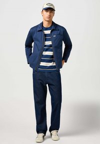 Chaqueta navy con cremallera, camiseta a rayas azul y blanca debajo, jeans azul oscuro y zapatillas blancas con acentos negros. Gorra con el logo de "Wrangler".