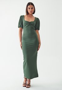 Zelena obleka s prilagojenim krojem in kvadratnim izrezom, nabranimi rokavi in nabranjem na prsi. Gladka tekstura, dolga do tal. Črne sandale z naramnicami.