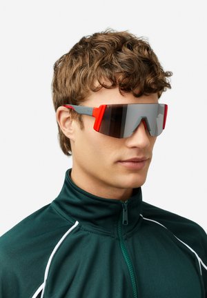 Päikeseprillid, millel on suured geomeetrilised läätsed hallide ja punaste aktsentidega. Õhukesed, sportlikud hallid raamid mattpinnaga.