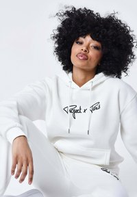 Set di felpa bianca con cappuccio e pantaloni abbinati, caratterizzato da logo ricamato nero, cappuccio con cordino e polsini a coste. Tessuto morbido con vestibilità comoda.