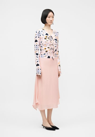 Femme aux cheveux courts noirs portant un haut à manches longues à motif floral, une jupe midi rose clair et des chaussures slingback noires à bout pointu.