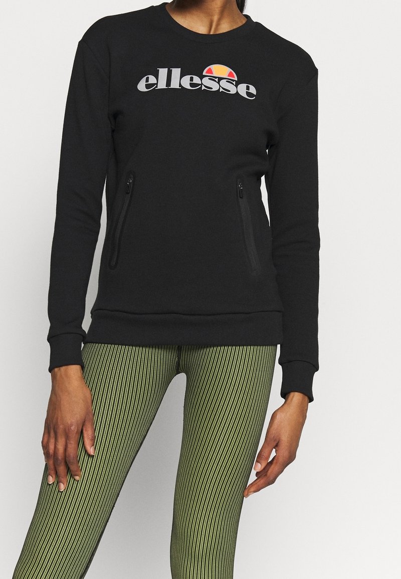 Czarna bluza z długimi rękawami, ściągaczami na mankietach, kieszeniami na zamek z przodu oraz szarym logo. Połączona z zielonymi legginsami w paski.