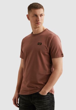 Junger Mann, der ein kurzärmliges T-Shirt in Rostfarbe und schwarze Jeans trägt, schaut zur Seite, eine Hand in der Tasche.