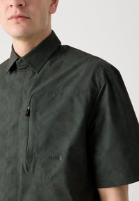 Korte mouwen shirt van donkergroene stof met een geometrisch patroon. Voorzien van een borstzak en een subtiele merkenlogo aan de zijkant.