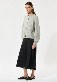 Lys grå strikk cardigan med rund hals, hulmønster og knaplukning, parret med en sort, flydende nederdel og spidse sorte flade sko.