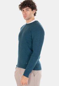 Maglione a maglia verde petrolio con motivo strutturato, collo rotondo e polsini a coste. Presenta un'etichetta con logo discreto sull'orlo. Indossato con pantaloni chiari.