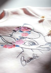 Tissu rose clair avec une illustration de Bambi comportant des motifs floraux en rose, bleu et des accents pailletés. Style à manches courtes et col rond.