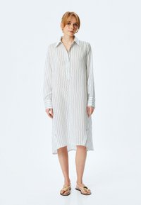 Abito a righe con bottoni in tessuto leggero, dotato di colletto, maniche lunghe, spacchi laterali e polsini a contrasto. Indossato con sandali flat.
