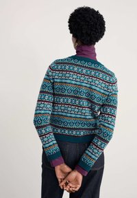 Adīta džemperis ar tirkīza fona krāsu, kurā ir daudzkrāsaini ģeometriski raksti, apaļa kakla izgriezums un ribotas piedurknes. Uzvilkts virs purpursarkanas turtlenecka.