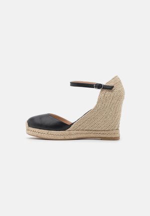 Buty espadryle na koturnie z czarnym noskiem, plecionym beżowym obcasem i czarnym paskiem wokół kostki z srebrną sprzączką na białym tle.