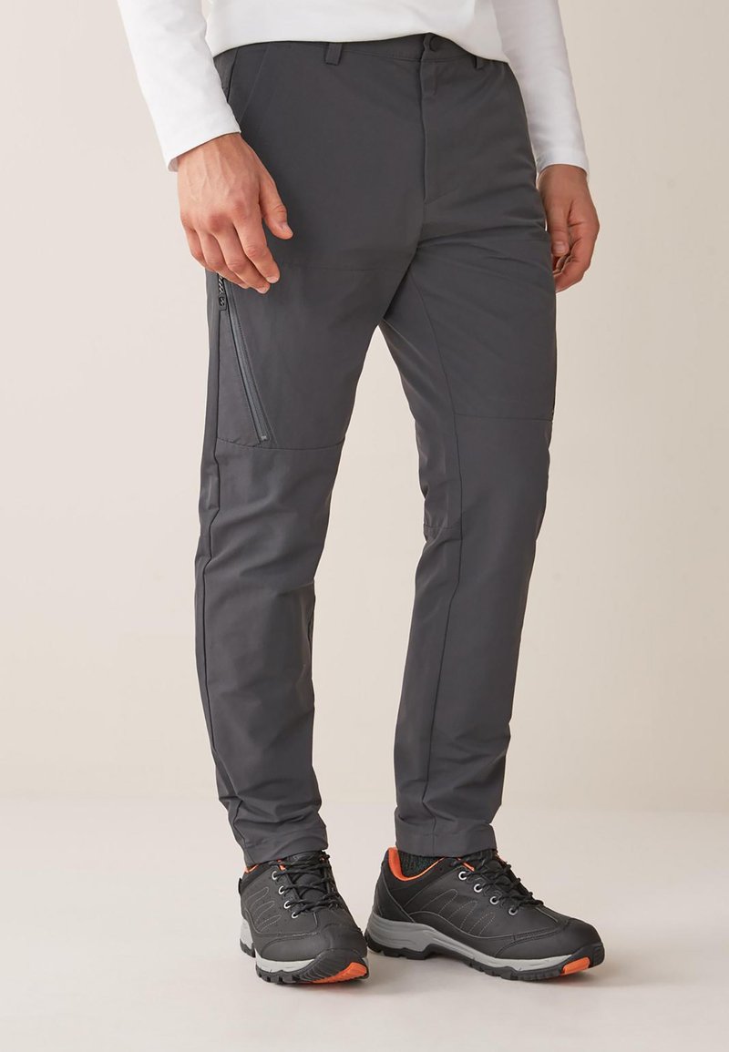 Pantaloni grigi, leggeri e resistenti all'acqua, con una tasca con zip, vestibilità affusolata e texture liscia. Abbinati a scarpe da trekking nere e arancioni.