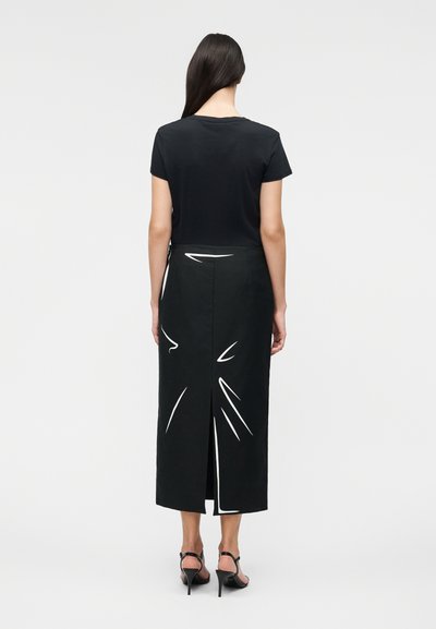 Γυναίκα με μακριά σκούρα μαλλιά που φοράει μαύρο τοπ με κοντά μανίκια και μαύρη μίντι φούστα με λευκές αφηρημένες γραμμές και σκίσιμο στην πλάτη, που στέκεται με την πλάτη γυρισμένη.