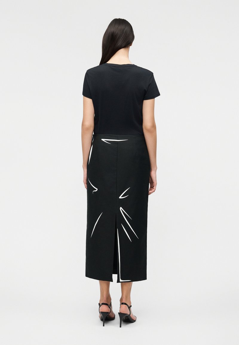 Woman with long dark hair wearing a black short-sleeve top and a black midi skirt with white abstract lines and back slit, standing facing away.