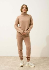 Beige neulepusero, jossa on korkea kaulus ja ribbireunat, yhdistettynä vastaaviin beigeihin housuihin. Mallilla on päällä valkoiset urheilukengät. Yksinkertainen, mukava muotoilu.