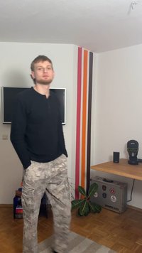 Zwart shirt met lange mouwen en beige camouflage cargobroek met zakken. Binnenomgeving met een houten tafel en een decoratieve plant.