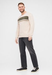 Suéter beige claro con rayas horizontales verdes oscuras y blancas, combinado con jeans gris oscuro y zapatillas negras, de pie sobre un fondo blanco.