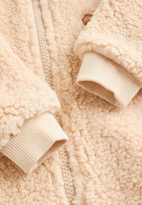 Light beige fleece jacket with a textured, fluffy exterior. Features a central zipper and ribbed cuffs for a snug fit.