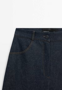Jeans en denim foncé avec une taille haute, une fermeture à boutons sur le devant et des coutures orange. Comprend des poches avant visibles et une texture lisse.