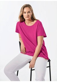 Magenta t-shirt met korte mouwen en een ronde hals, gemaakt van zachte stof. Gekozen met witte broeken, zittend op een zwarte stoel.