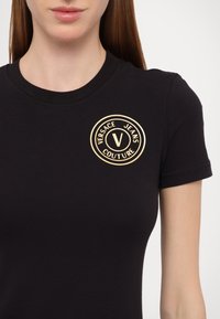 Crna pamučna majica s uskim krojem, kratkim rukavima i zlatnim okruglim logom koji sadrži "Versace Jeans Couture" na prsima.