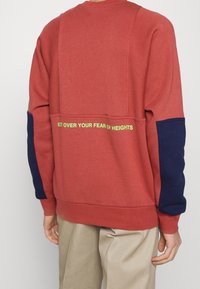 Röd sweatshirt med marinblå detaljer, med en stor bakpanel och texten "ÖVERVINNER DIN RÄDSLA FÖR HÖJDER" i neongrön.