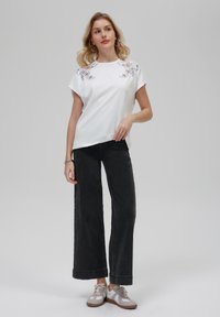 T-shirt blanc à manches courtes avec broderie florale au niveau des épaules, associé à un jean large noir et des baskets grises.