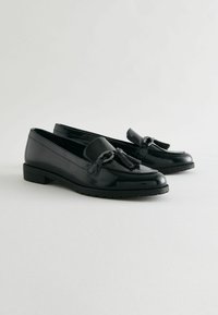 Loafers din piele patent neagră cu o talpă plată, având un ciucure și un detaliu de lanț decorativ pe față. Finisaj neted și lucios.