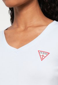 Zbliżenie na osobę w jasnoniebieskiej koszulce z dekoltem w serek z czerwono-białym logo Guess na górnej części klatki piersiowej.