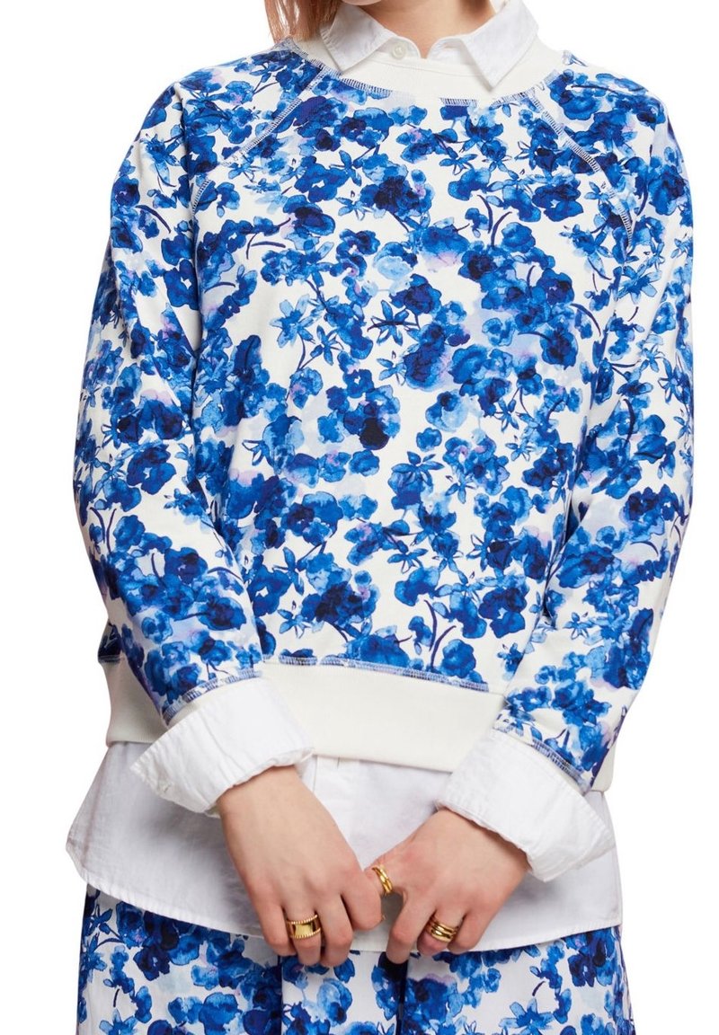 Blommig sweatshirt med blå och vita mönster, tillverkad av mjukt material. Rund halsringning med ribbade mudd och nederkant. Lagd över en vit skjorta.