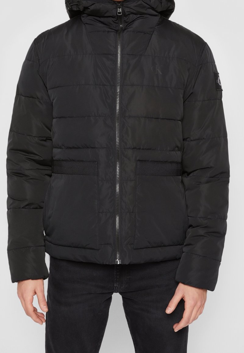 Svart pufferjacka med huva, med quiltat mönster, framdragkedja och två stora sidofickor. Texturerat tyg med elastiska detaljer.