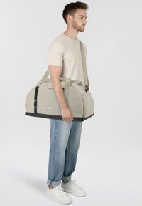Sac de sport gris clair avec une base noire, doté de poignées doubles, d'une sangle d'épaule réglable et d'une poche latérale en mesh. Fini en tissu texturé.