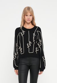 Črni pleteni pulover z oversized krojem, ki ima bele abstraktne linijske vzorce. Okrogli izrez in dolgi rokavi. Teksturirana tkanina.