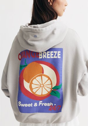 Hoodie oversize gris avec un graphique coloré de fruits au dos, accompagné du texte "Orange Breeze" et "Sweet & Fresh 24/7". Tissu doux.