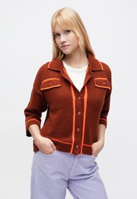 Pruun kootud kardigan oranži äärisega, kahe esitasku ja nööpidega sulguriga. Omab kraed ja kolmveerand pikkusega varrukaid. Tekstureeritud kangas.