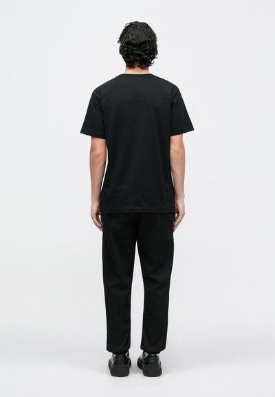 Čierne bavlnené tričko s krátkymi rukávmi, rovný strih, nosené s čiernymi nohavicami a topánkami, zobrazené zozadu na jednoduchom pozadí.