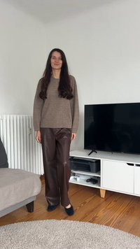 Pulover maro tricotat, asortat cu pantaloni largi maro din piele ecologică. Pantofi slip-on negri și fundal neutru cu mobilier.
