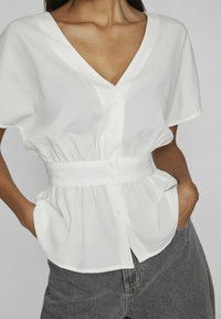 Blusa bianca con scollo a V, maniche corte e vita stretta. Presenta bottoni sul davanti e due tasche laterali. Tessuto leggero.