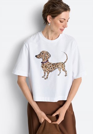 Frau trägt weißes kurz geschnittenes T-Shirt mit einem glitzernden gefleckten Dackel-Design, braunen Rock und silberne Ohrringe, hält braunen Stoff.