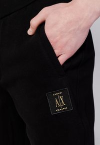Pantaloni jogger neri con una patch del logo Armani Exchange in oro, caratterizzati da una tasca laterale e una trama di tessuto morbido.