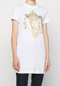 Biała bawełniana koszulka z krótkim rękawem, z dużym złotym nadrukiem graficznym okrągłego emblematu i dekoracyjnych elementów na środku.