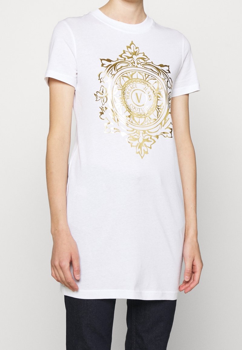Biała bawełniana koszulka z krótkim rękawem, z dużym złotym nadrukiem graficznym okrągłego emblematu i dekoracyjnych elementów na środku.
