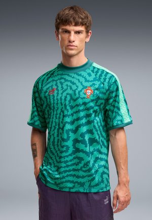Muž nosiaci zelený futbalový dres Portugalska s abstraktnými vzormi a fialové Puma šortky, stojaci na sivom pozadí.