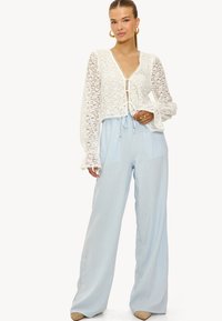 Haut en dentelle blanche à manches longues avec un décolleté en V et une fermeture à nouer, associé à un pantalon bleu clair à jambes larges. Les deux vêtements présentent des tissus fluides.