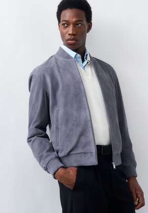 Άνδρας που φοράει ελαφρύ γκρίζο σουέντ μπουφάν bomber, λευκό πουλόβερ, γαλάζιο πουκάμισο με γιακά και μαύρο παντελόνι, στέκεται με το ένα χέρι στην τσέπη.