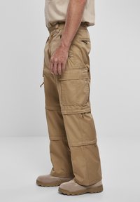 Beige cargo-byxor har flera fickor, en lös passform och dragkedjor. Designade för funktionalitet med ett hållbart tyg.